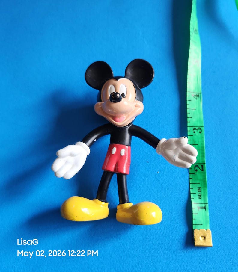 May include: A Mickey Mouse figurine with black ears, a black body, red shorts, yellow shoes, and white gloves. A green measuring tape is next to the figurine. The figurine is approximately 3 inches tall.