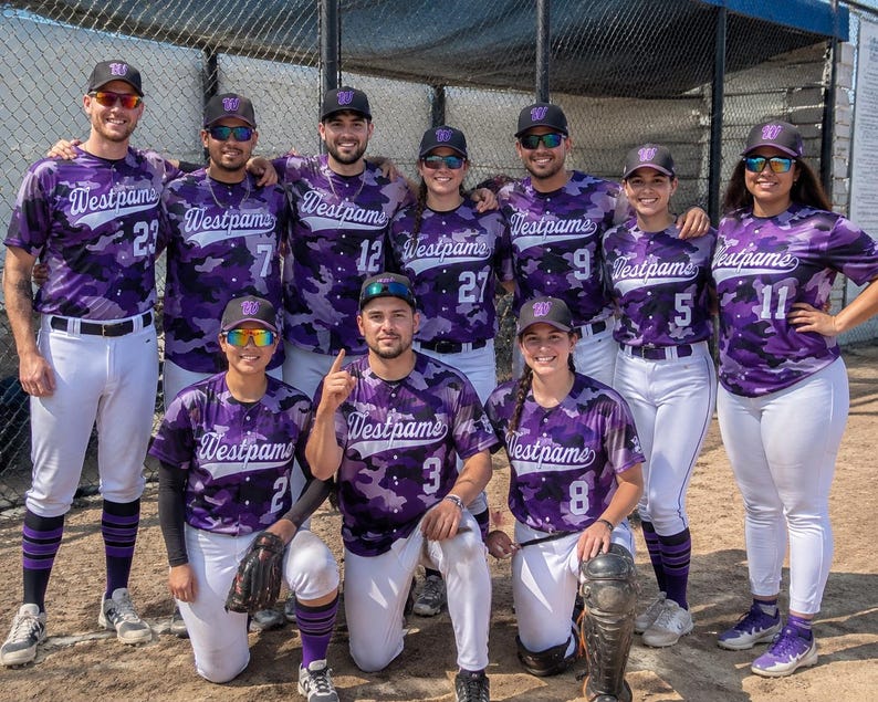 May include: A group of ten baseball players wearing matching purple and black camouflage jerseys with the text Westpams printed on the front. Each player wears white pants, a purple baseball cap, and sunglasses while posing on a dirt field.