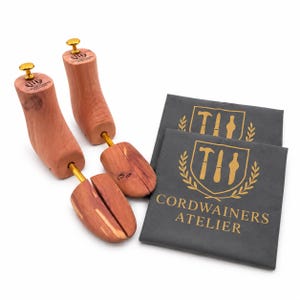 May include: A pair of light brown wooden shoe trees with gold-toned metal knobs and hardware, accompanied by two dark grey fabric pouches featuring a gold logo with the text CORDWAINERS ATELIER and an illustration of a hammer, knife, and tool.