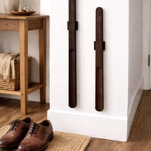 May include: Two dark brown wooden shoe racks fixed to a white wall. The racks are rectangular with a curved top and a circular detail. Brown leather shoes with black laces sit on a woven mat on the floor.