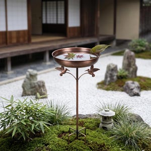 May include: A bronze bird bath with a shallow bowl, supported by a slender stand and decorative bird accents. The bowl contains water and fallen leaves. A small green bird is perched on the edge, drinking. The bird bath is set in a Japanese garden.