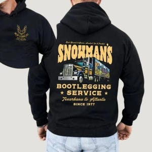 May include: Black hoodie with a graphic on the back that reads "Snowman's Bootlegging Service" with a truck illustration. The hoodie also features text that says "Texarkana to Atlanta Since 1977". The left shoulder has a gold graphic.