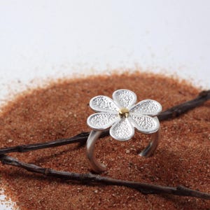May include: A silver flower ring with a gold center, set against a backdrop of brown sand and a twig. The ring's flower design features textured petals, creating a delicate and nature-inspired aesthetic.