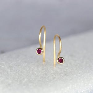 Peut inclure: Une paire de boucles d'oreilles pendantes dorées. Chaque boucle d'oreille présente une petite pierre précieuse ronde, rouge foncé, sertie dans une lunette dorée. Les boucles d'oreilles ont un design simple et incurvé, avec la pierre précieuse en bas.