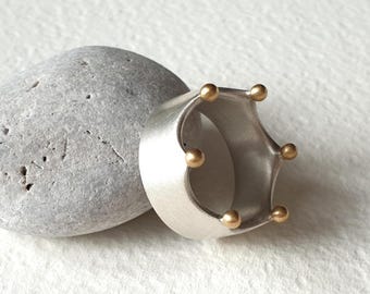 May include: A silver-colored ring with a crown design, featuring six small gold-colored spheres. The ring is placed next to a gray stone on a white surface. The ring's design is modern and minimalist.