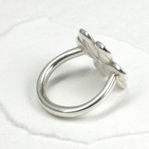 May include: A silver ring featuring a flower design. The ring has a thick band and a detailed floral element with multiple petals. The ring is shown on a white surface.