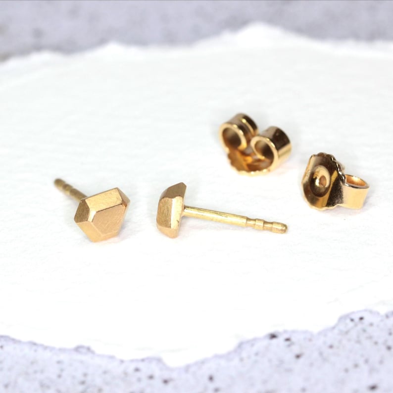 Op de afbeelding: Goudkleurige oorstekers met een geometrisch ontwerp. De oorbellen hebben een facetvormige, diamantachtige vorm. Twee oorbelruggen zijn ook zichtbaar. De oorbellen worden gepresenteerd op een wit oppervlak.