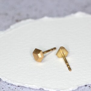 Op de afbeelding: Twee goudkleurige oorstekers. Elke oorbel heeft een geometrisch, facetontwerp. De oorbellen worden gepresenteerd op een wit, getextureerd oppervlak. De pinnen zijn dun en goudkleurig.