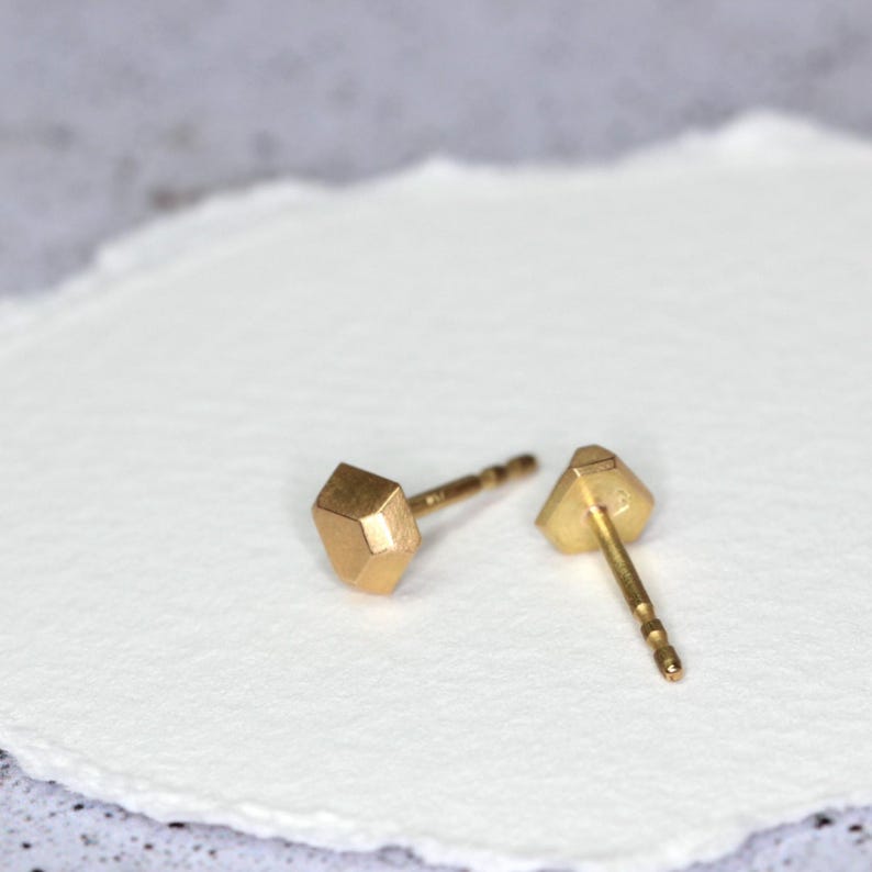 Op de afbeelding: Twee goudkleurige oorstekers. Elke oorbel heeft een geometrisch, facetontwerp. De oorbellen worden gepresenteerd op een wit, getextureerd oppervlak. De pinnen zijn dun en goudkleurig.