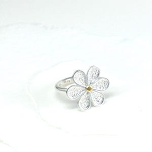 May include: A silver ring featuring a daisy flower design. The flower has five textured petals and a small gold-colored center. The ring is set against a white background, highlighting the jewelry's details.