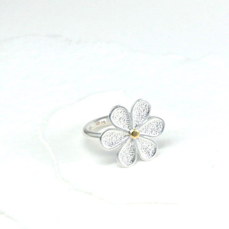 May include: A silver ring featuring a daisy flower design. The flower has five textured petals and a small gold-colored center. The ring is set against a white background, highlighting the jewelry's details.