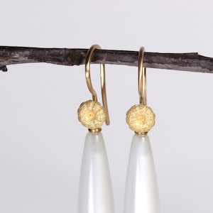 May include: Gold-coloured drop earrings featuring a textured, round top and a white, teardrop-shaped pearl. The earrings are suspended from a thin, dark brown branch against a white backdrop.