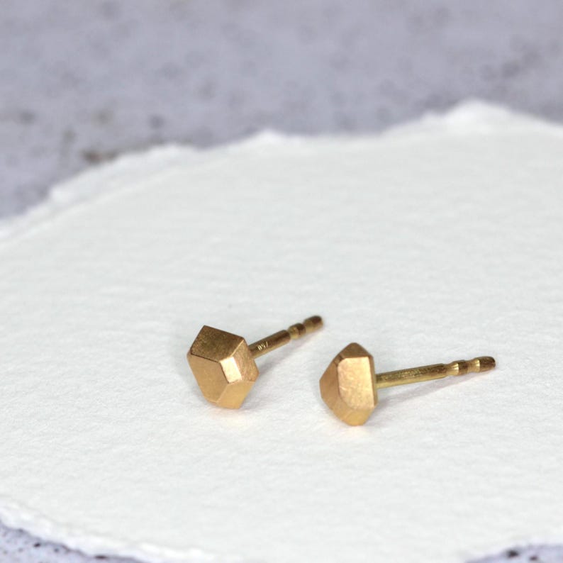 Op de afbeelding: Een paar kleine, goudkleurige oorstekers. Elke oorbel heeft een geometrisch, facetgeslepen ontwerp. De oorbellen worden gepresenteerd op een wit, getextureerd oppervlak, met een zachte, vage achtergrond.
