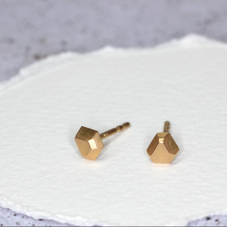 Op de afbeelding: Een paar kleine, goudkleurige oorstekers. Elke oorbel heeft een geometrisch, facetgeslepen ontwerp. De oorbellen worden gepresenteerd op een wit oppervlak, met een zachte, vage achtergrond.