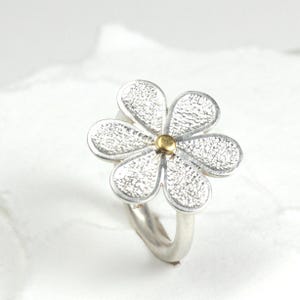May include: A silver flower ring with a textured surface on the petals and a gold-colored center. The ring has a simple, curved band. The flower design is the focal point of this jewelry piece.
