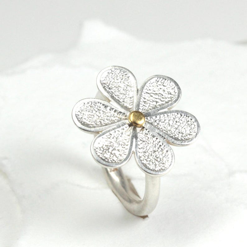 May include: A silver flower ring with a textured surface on the petals and a gold-colored center. The ring has a simple, curved band. The flower design is the focal point of this jewelry piece.