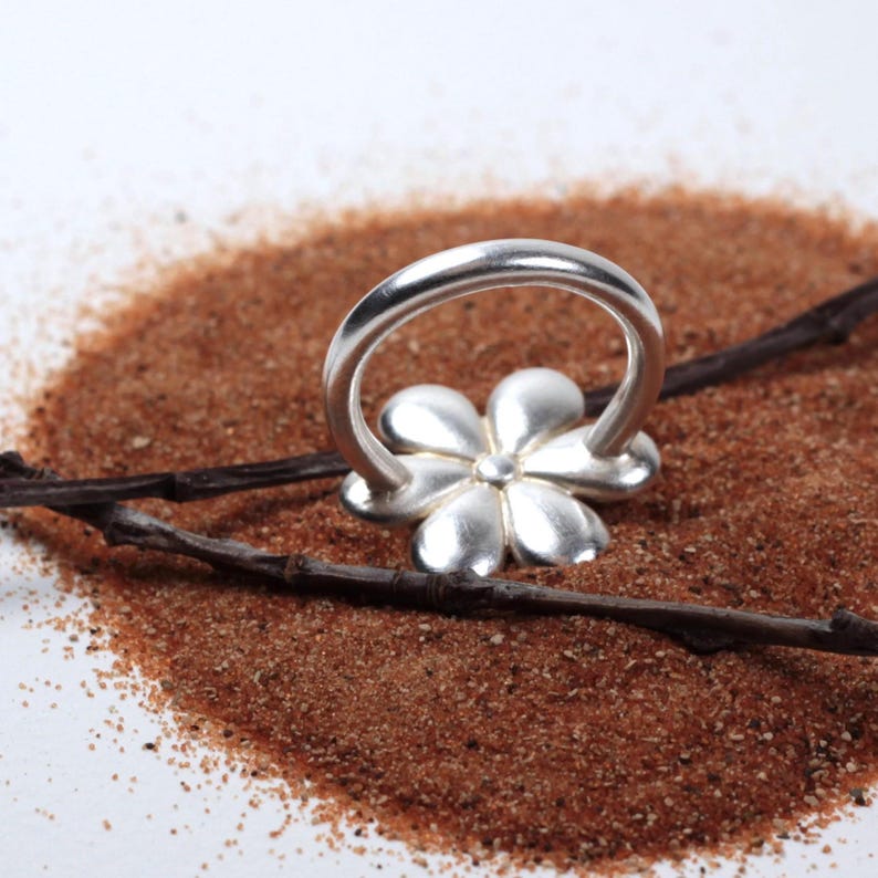 May include: A silver-colored ring with a flower design. The ring features a floral motif with five petals and a smooth, polished band. The ring is set against a backdrop of brown sand and a dark twig.