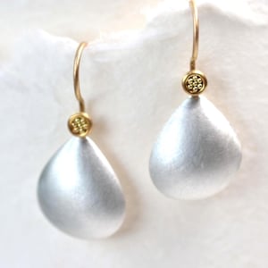 Peut inclure: Paire de boucles d'oreilles argentées en forme de goutte d'eau, avec des crochets d'oreille dorés et de petits accents ronds dorés. Les boucles d'oreilles ont une finition en métal brossé et un design simple et élégant.