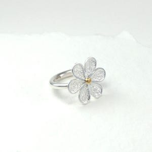 May include: A silver ring featuring a daisy flower design. The flower has five petals with a textured, glittery appearance and a small gold-colored center. The ring band is smooth and silver.