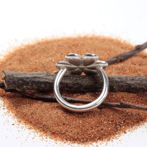 May include: A silver ring with a four-petal flower design. The ring is set against a backdrop of sand and a small twig. The flower petals are rounded and smooth, and the band is a simple, polished silver.