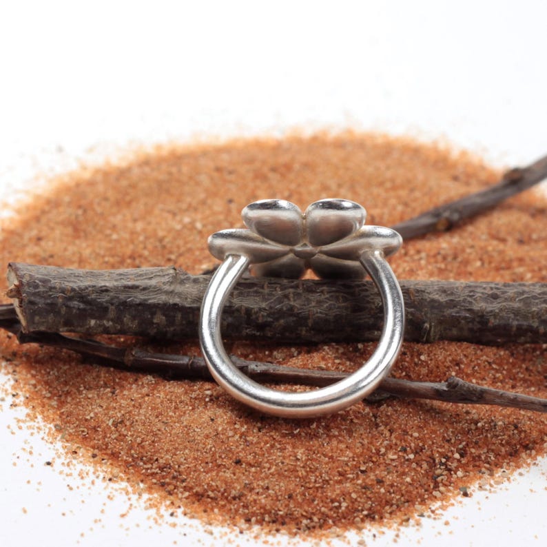 May include: A silver ring with a four-petal flower design. The ring is set against a backdrop of sand and a small twig. The flower petals are rounded and smooth, and the band is a simple, polished silver.