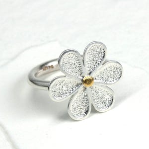 May include: A silver ring featuring a daisy flower design. The flower has textured petals and a gold-colored center. The ring is set against a white background.