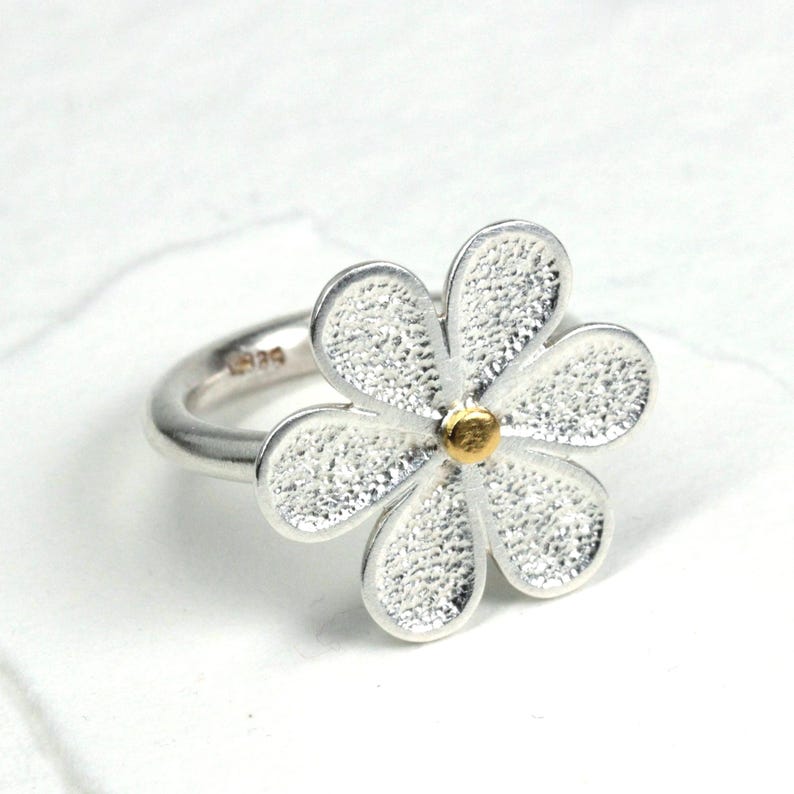 May include: A silver ring featuring a daisy flower design. The flower has textured petals and a gold-colored center. The ring is set against a white background.