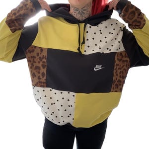 Può includere: Una felpa con cappuccio patchwork con cappuccio e maniche nere, con pannelli gialli, neri, bianchi a pois neri e stampa leopardata. Il logo Nike è visibile sul davanti.
