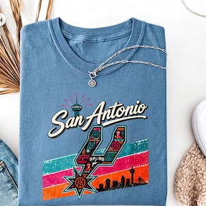 May include: A blue crew neck t-shirt featuring a graphic print that says San Antonio in script font. The design includes a stylised number four, a basketball, a city skyline, and colourful horizontal stripes in teal, pink, and orange hues.