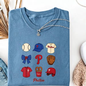 Może przedstawiać: Niebieski t-shirt z okrągłym dekoltem z nadrukiem graficznym przedstawiającym ikony baseballowe, w tym piłkę, niebieską czapkę, koszulkę w paski, niebieską kokardę, czerwoną literę P, brązową rękawicę, czerwoną koszulkę, precel i czerwony kask.