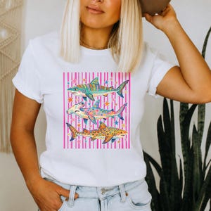 Può includere: Una t-shirt bianca a maniche corte con una stampa grafica di tre squali impilati verticalmente. Gli squali sono decorati con motivi colorati e complessi su uno sfondo di strisce verticali rosa e piccole stelle multicolori.