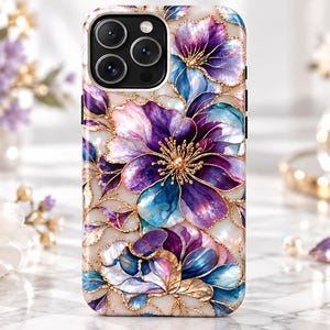 Könnte beinhalten: Eine Smartphone-Hülle mit einem lebendigen Blumenmuster aus dunkelvioletten, blauen und weißen Blütenblättern. Das Design zeigt filigrane goldfarbene Umrandungen um jede Blume und jedes Blatt auf einem perlmuttfarbenen, hellen Hintergrund.