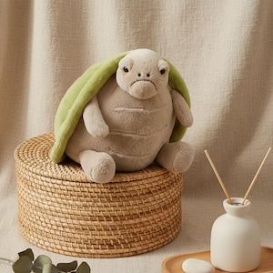 Puede incluir: Un peluche de tortuga con caparazón verde claro y cuerpo beige sentado sobre una cesta tejida. La tortuga tiene una expresión ligeramente gruñona. El fondo incluye un difusor de cerámica blanca y una bandeja de madera.