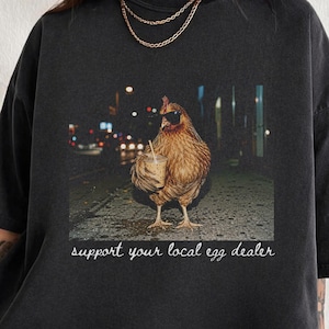 May include: A black short-sleeve t-shirt featuring a graphic of a brown chicken wearing sunglasses and holding a plastic cup with a straw. Below the chicken, the white text reads, support your local egg dealer. The shirt has a crew neckline.