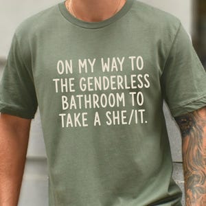 May include: An olive green short-sleeved t-shirt featuring white text that reads: ON MY WAY TO THE GENDERLESS BATHROOM TO TAKE A SHE/IT. The shirt has a crew neckline and a standard fit, displaying the printed message clearly across the chest area.