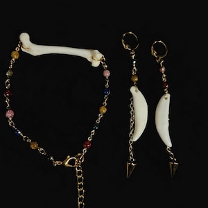 May include: A jewelry set consisting of a bracelet and two earrings. The bracelet features a small white bone segment connected to a gold chain with multicolored beads. The earrings feature gold hoops, small beads, white curved bone pendants, and gold spikes.