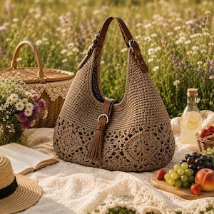 Op de afbeelding: Een beige gehaakte hobo-tas met een bloemmotief, bruine leren banden met zilveren gespen en een centrale leren band met een bijpassend kwastje. De tas is gemaakt van gevlochten koord en heeft een gestructureerd ontwerp met handvat.