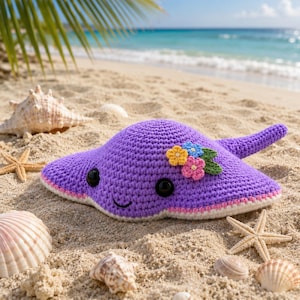 May include: A handmade purple crochet stingray with a smiling face and flower accents, resting on a sandy beach. The stingray has black button eyes, a pink and white border, and is surrounded by seashells and starfish.