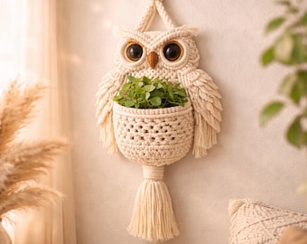 May include: A cream-colored macrame owl-shaped hanging planter with a woven basket for plants. The owl has large, dark eyes and a wooden beak. The wings and a tassel at the bottom add texture. The planter is filled with green plants.