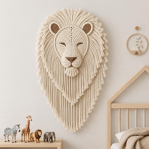 May include: A wall hanging crafted from off-white macrame cord, shaped like a lion head with a full, layered mane of fringe. The lion features brown accents on the ears, eyes, and nose, with a textured, knotted face and a small tufted chin.