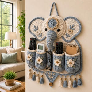 Peut inclure: Un organisateur mural en crochet en forme d'éléphant aux tons bleu et crème. Il comporte quatre poches cylindriques décorées de fleurs couleur crème et de boutons en bois, ainsi qu'un espace central pour des lunettes et des pompons en bas.