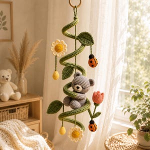 Op de afbeelding: Handgemaakte gehaakte baby mobile met een grijze beer genesteld in een groene wijnstok. De mobiel bevat gele bloemen, oranje lieveheersbeestjes en een roze tulp. De mobiel is opgehangen aan een houten ring.