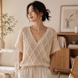 May include: A cream-coloured, short-sleeved V-neck sweater with a diamond-patterned knit. The sweater is worn with light-coloured trousers. The overall aesthetic is casual and comfortable, suitable for everyday wear. The image is well-lit, highlighting the texture of the knit.