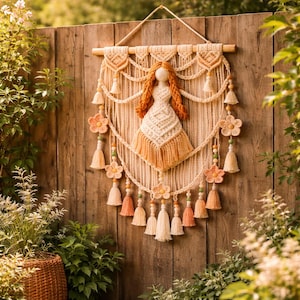May include: A decorative macrame wall hanging with a doll-like figure. It features orange hair and a cream dress. The design includes tassels, flower accents, and a wooden rod. The colour scheme is cream, orange, and peach, suitable for a bohemian-style interior.