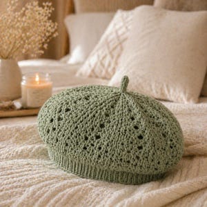 May include: A sage green crochet beret with a textured, openwork design. The beret features a ribbed brim and a small, decorative loop at the top. The hat is displayed on a cream-colored surface.