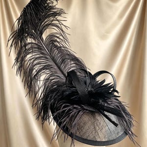 May include: A black fascinator hat featuring a large, upright ostrich feather plume, a central fabric flower, and two curved ribbon loops. The base is constructed from a fine, woven mesh material with a solid black trim along the outer edge.