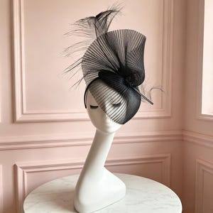 May include: A black fascinator hat featuring a structured, pleated mesh base that curves upward. The headpiece is adorned with long, delicate black feathers extending from the top and a small, dark circular embellishment at the center of the design.