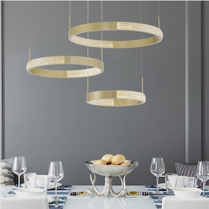 Könnte beinhalten: Drei goldfarbene, kreisförmige Pendelleuchten hängen über einem Esstisch. Jede Leuchte hat einen Abschnitt mit eingebetteten, funkelnden Kristallen. Der Tisch ist mit Glaswaren, Tellern und einer dekorativen Schale mit Brötchen gedeckt.