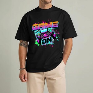 May include: Black t-shirt featuring a colorful graphic of a video game controller with the words "GAME ON". The design includes neon green, purple, blue, and orange accents, creating a vibrant and eye-catching look.
