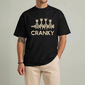 May include: Black t-shirt with a beige graphic of an engine crankshaft and the word "CRANKY" in a distressed font. The shirt is a crew neck and has short sleeves. The model is wearing khaki pants.